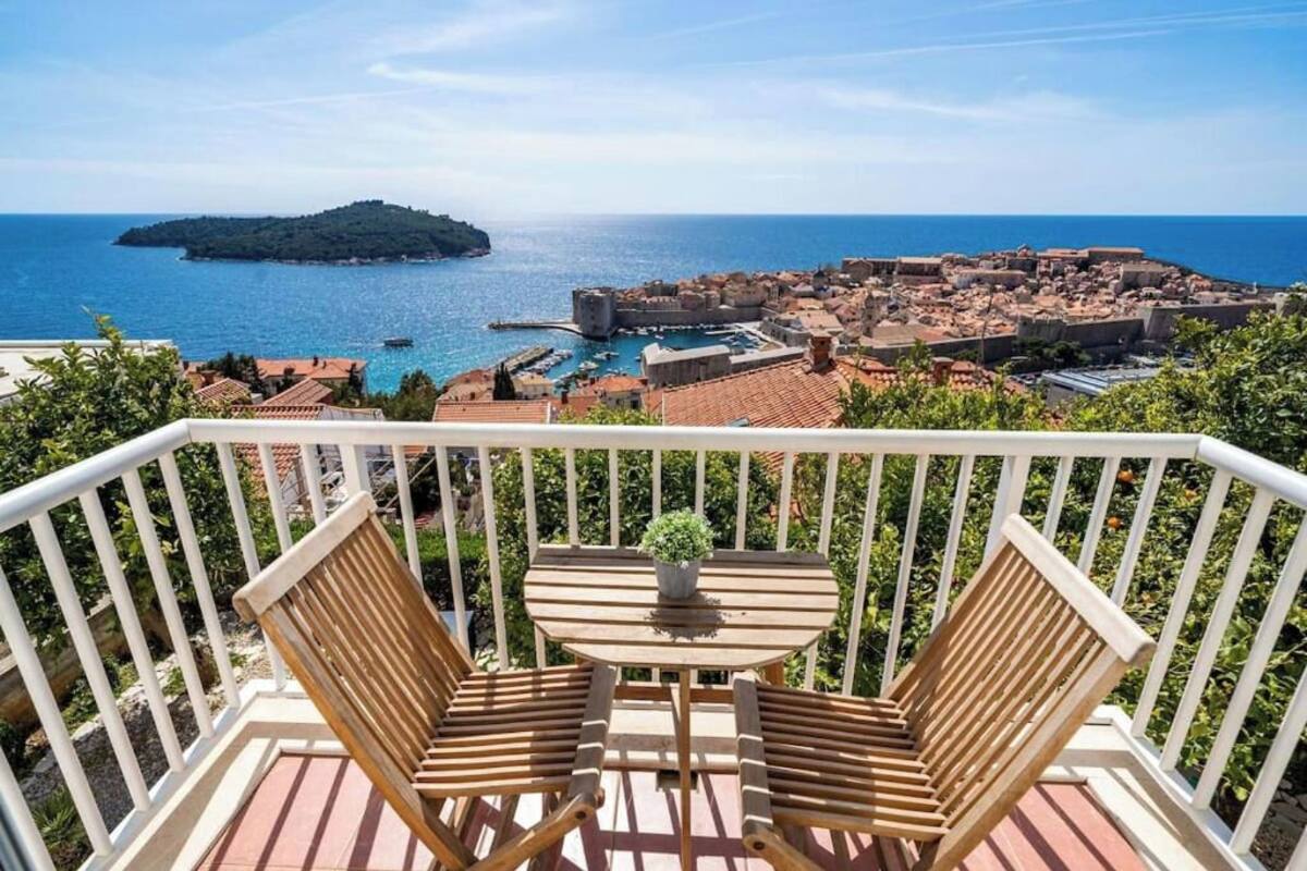 En balkon med to træstole og et lille bord, med udsigt over en kystby med en historisk fæstning og en lille ø i horisonten.