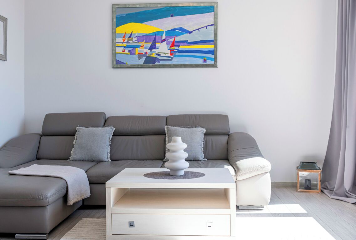 Un salón moderno con un sofá gris, una mesa de centro blanca con un jarrón decorativo, y un cuadro de veleros en la pared.