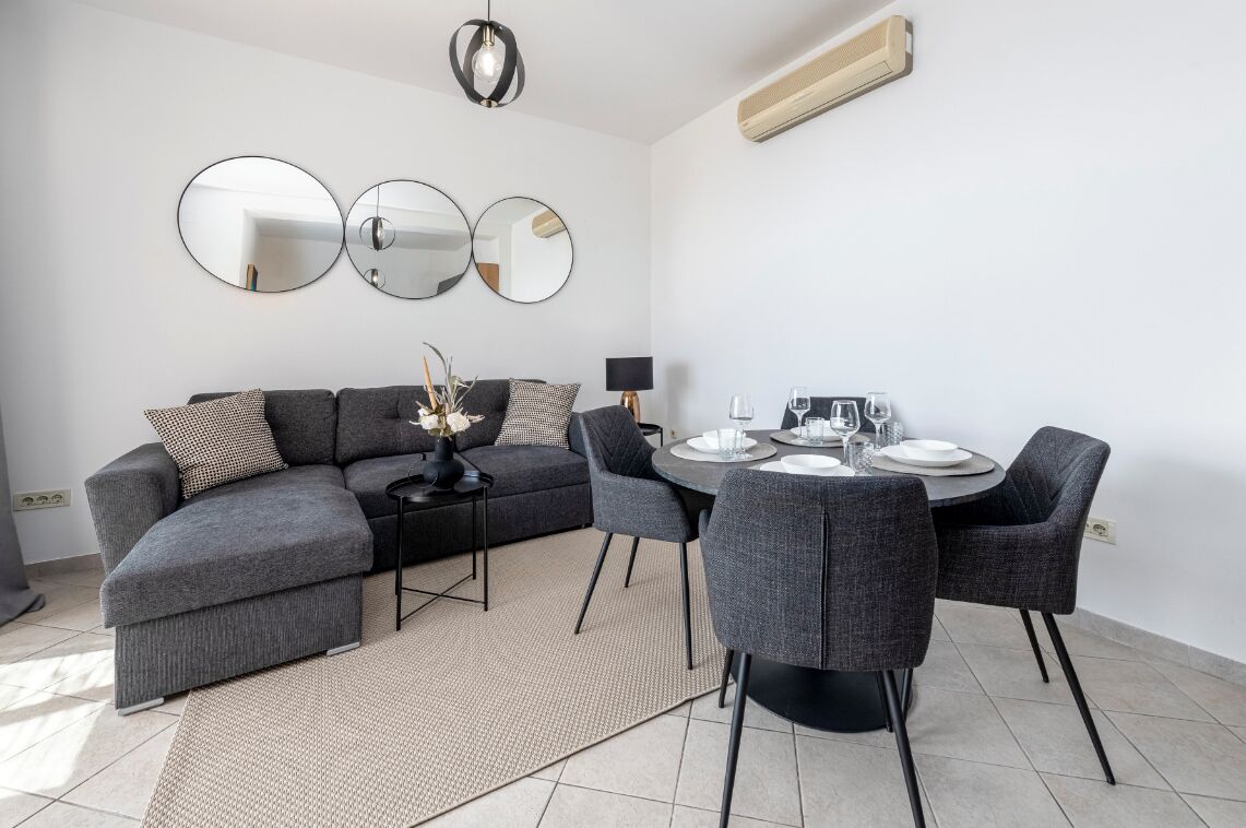 Un salon moderne avec un canapé d’angle gris, une table de salle à manger ronde entourée de quatre chaises et trois miroirs fixés au mur.