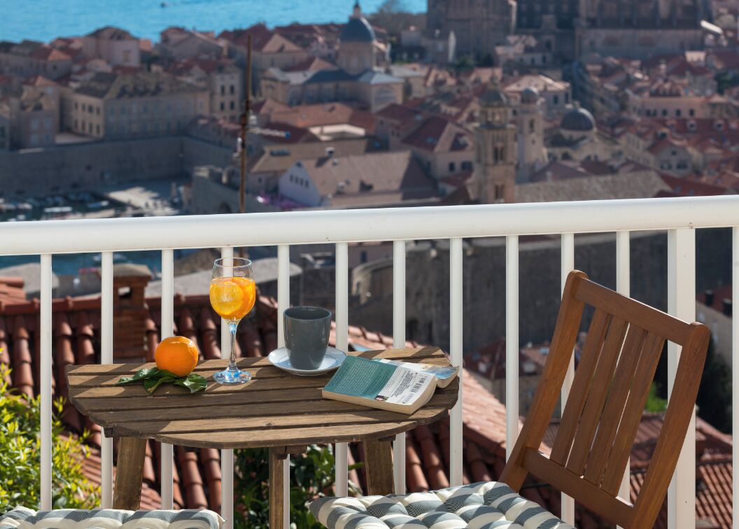 A wooden table with a book, a glass of orange juice, and a cup on a balcony overlooking a cityscape.