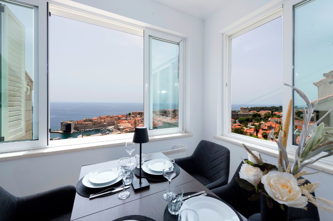 A dining area with a table set for two, large windows with a view of a coastal town and the sea.
