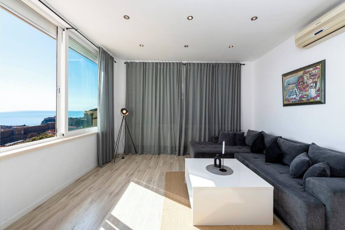 En moderne stue med et stort vindue, grå gardiner, en grå sofa, et hvidt sofabord og en gulvlampe.