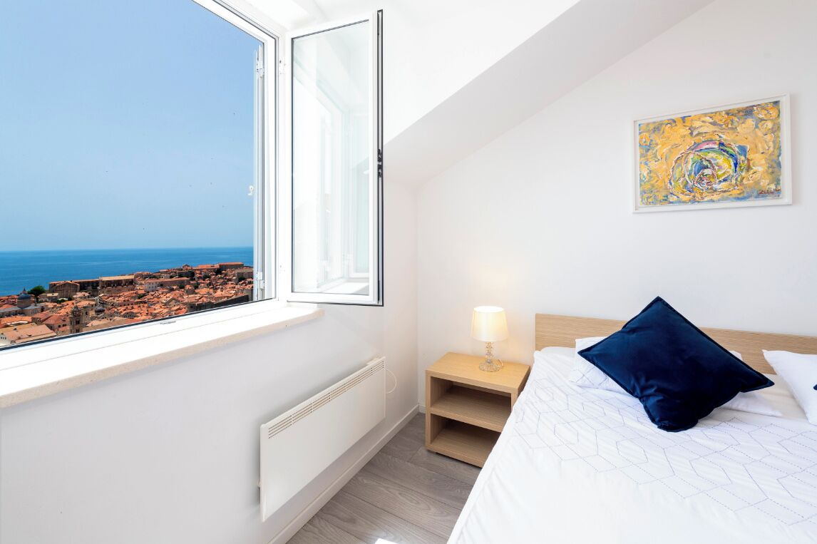 A bedroom with a large window, a bed with a blue pillow, a bedside table with a lamp, and a painting on the wall.