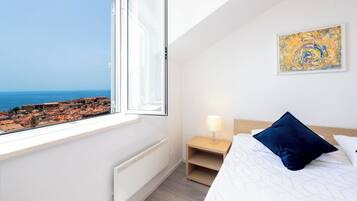 A bedroom with a large window, a bed with a blue pillow, a bedside table with a lamp, and a painting on the wall.