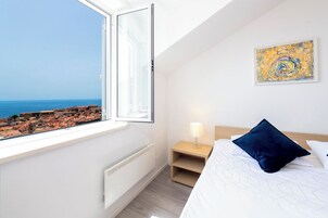 Quarto com uma janela grande, uma cama com travesseiro azul, mesa de cabeceira com abajur e um quadro na parede.