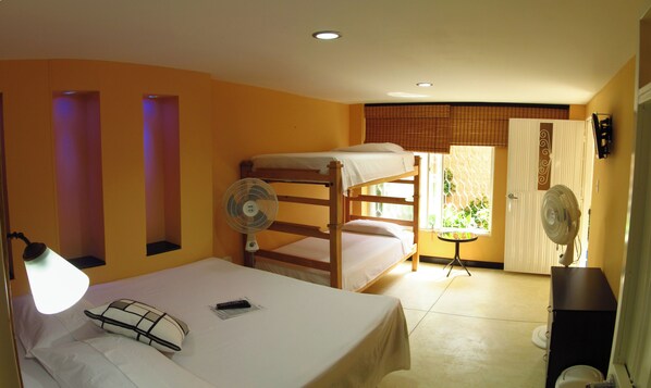 A room with bunk beds, a single bed, a small table, two fans, and a window with a view of greenery. - Hotel Casa Dmer (Taganga)