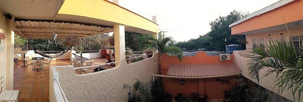 A covered outdoor seating area with tables and chairs, surrounded by potted plants and a view of a residential area. - Hotel Casa Dmer (Taganga)