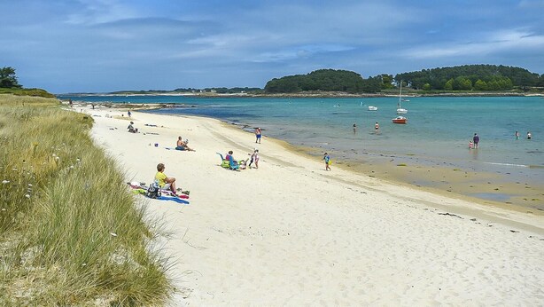 Ein Strand mit Menschen, die sich sonnen und schwimmen, Booten im Wasser und einer grünen Fläche links.