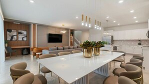 A modern living room with a large white table, comfortable seating, a flat-screen TV, and a kitchen area with white cabinets and a refrigerator. - Incredible 10bd Summerkitchen Windsor Cay (Clermont)