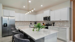 A modern kitchen with white cabinets, stainless steel appliances, a large island with bar stools, and a hanging light fixture. - Incredible 10bd Summerkitchen Windsor Cay (Clermont)
