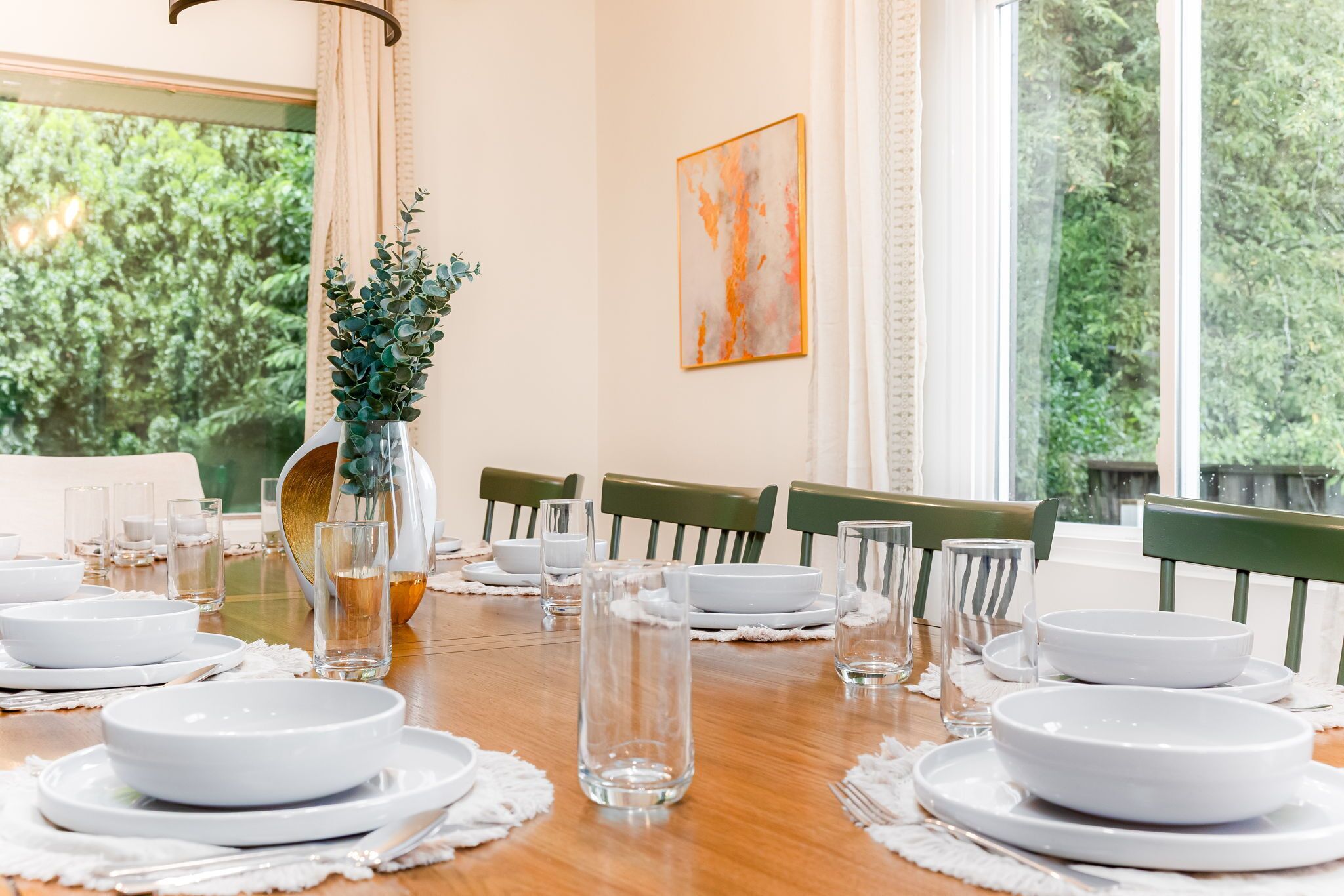 A dining table set for six with white plates, clear glasses, and a vase with greenery.