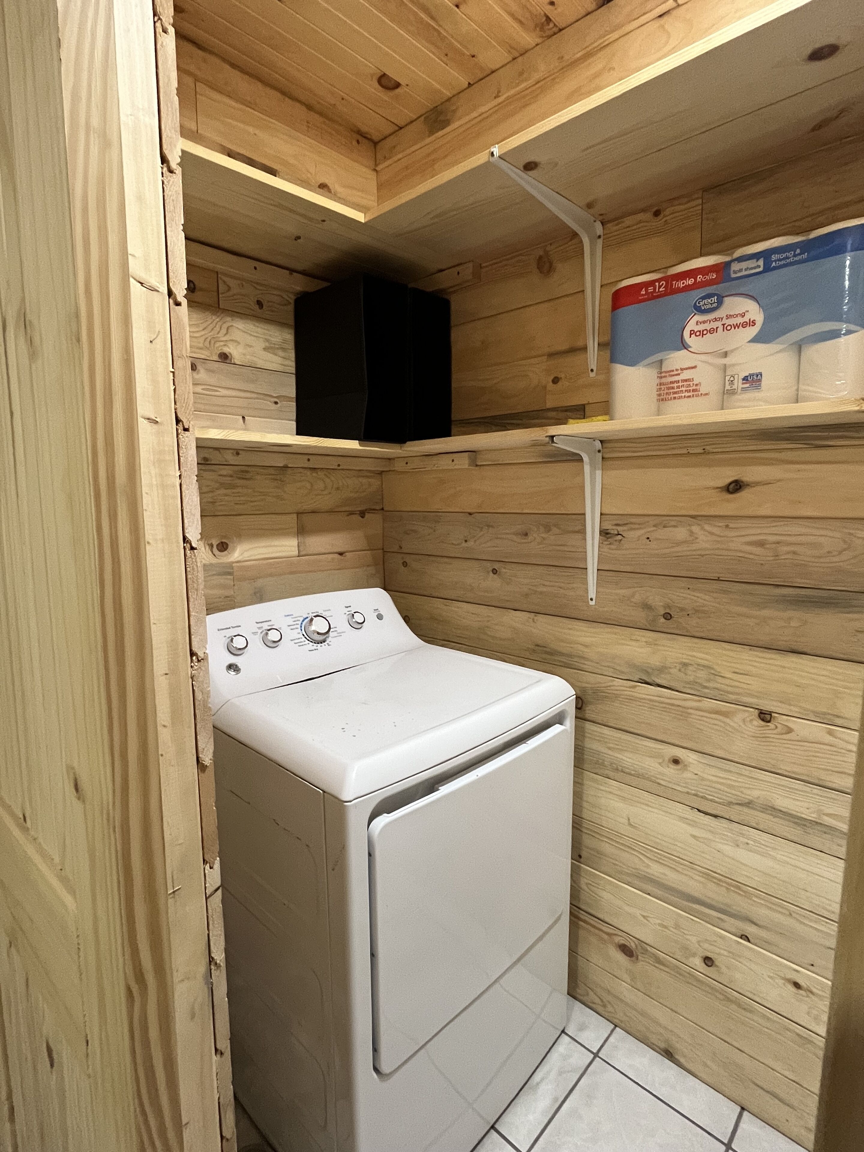 Un lavadero pequeño con lavadora y secadora blancas, paredes de madera y estantes con toallas de papel.