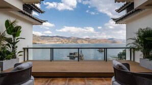 Un balcón con vistas a un lago y montañas, con mobiliario de mimbre y plantas en macetas.