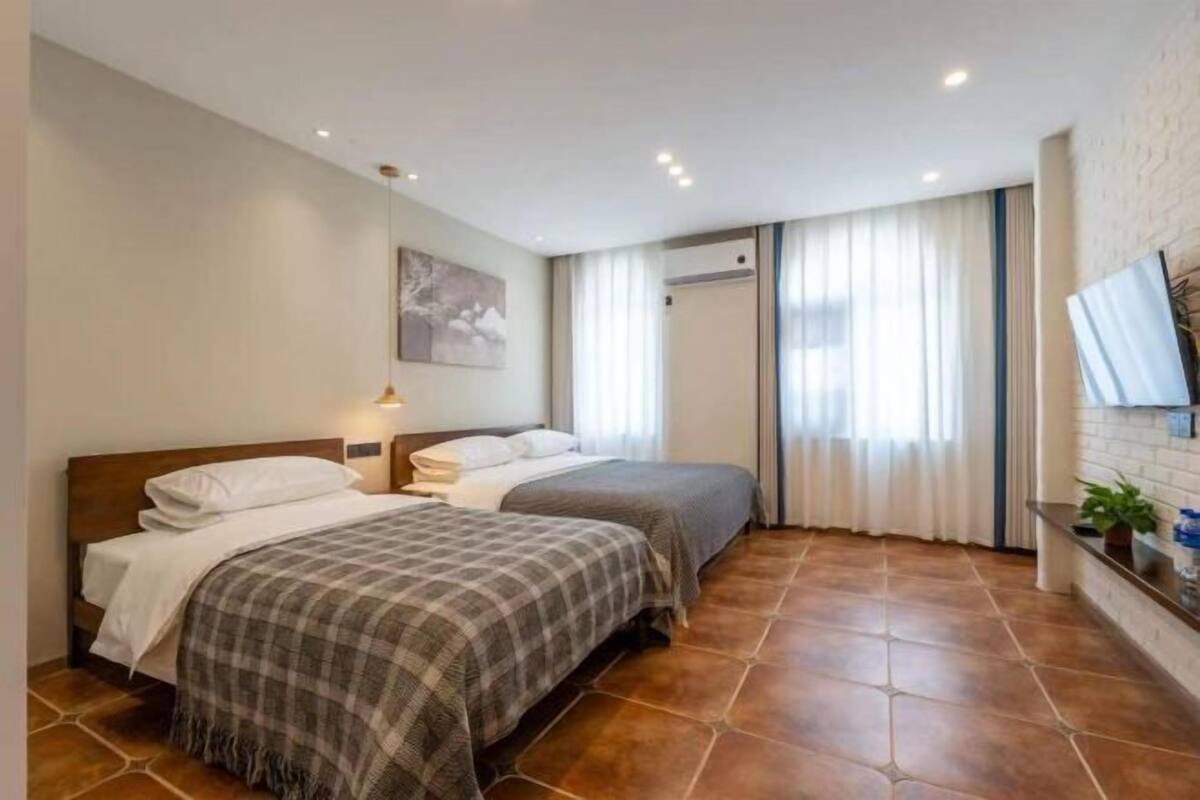 A hotel room with two beds, a large window with curtains, a flat-screen TV mounted on the wall, and a tiled floor.