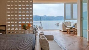 Una habitación de hotel moderna con una cama grande, una zona de estar con sofá y sillones, una mesa de comedor y vistas al mar a través de amplios ventanales.