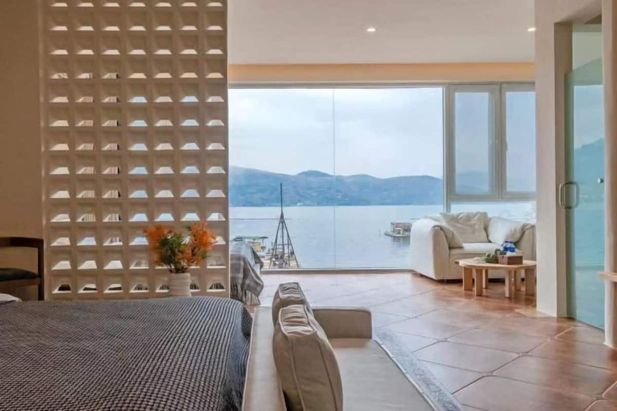 A modern hotel room with a large bed, a seating area with a sofa and armchairs, a dining table, and a view of the sea through large windows.