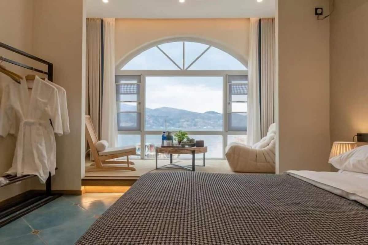 A bedroom with a large bed, a seating area with a table, a view of the outdoors, and a window with curtains.