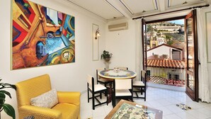 Een kamer met een gele bank, een ronde tafel met een decoratief bord, een kleurrijk abstract schilderij en een uitzicht op gebouwen door een open deur.