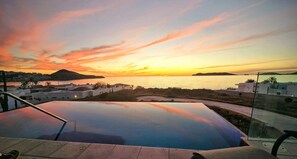 Una piscina infinita con vistas a la puesta de sol sobre el mar y un pueblo costero.