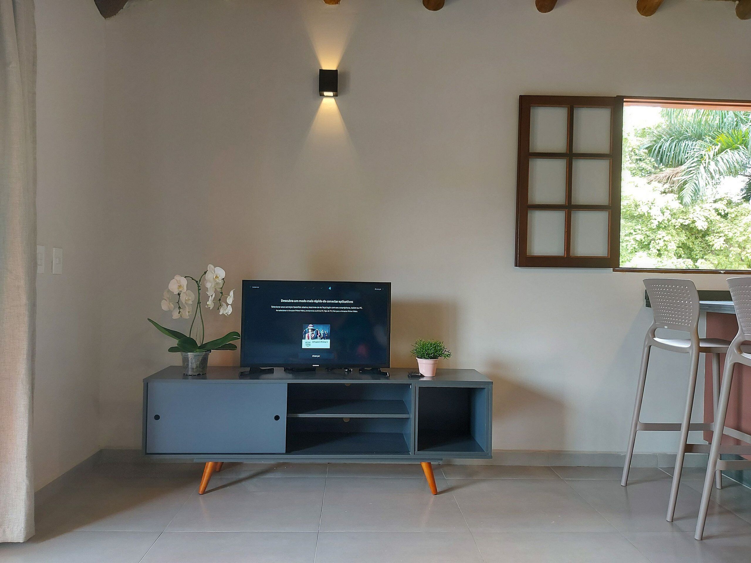 A modern living room with a flat-screen TV on a grey entertainment unit, a small potted plant, and a window with a view of greenery.