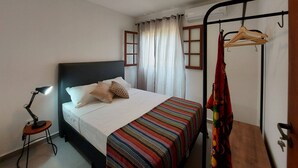 A bedroom with a bed, a bedside table, a hanging light, a window with curtains, and a coat rack with a coat.