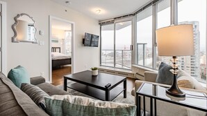 Ein modernes Wohnzimmer mit einer Couch, einem Couchtisch und einer Lampe. Ein Fernseher ist an der Wand montiert und es gibt ein Fenster mit Blick auf die Stadt.