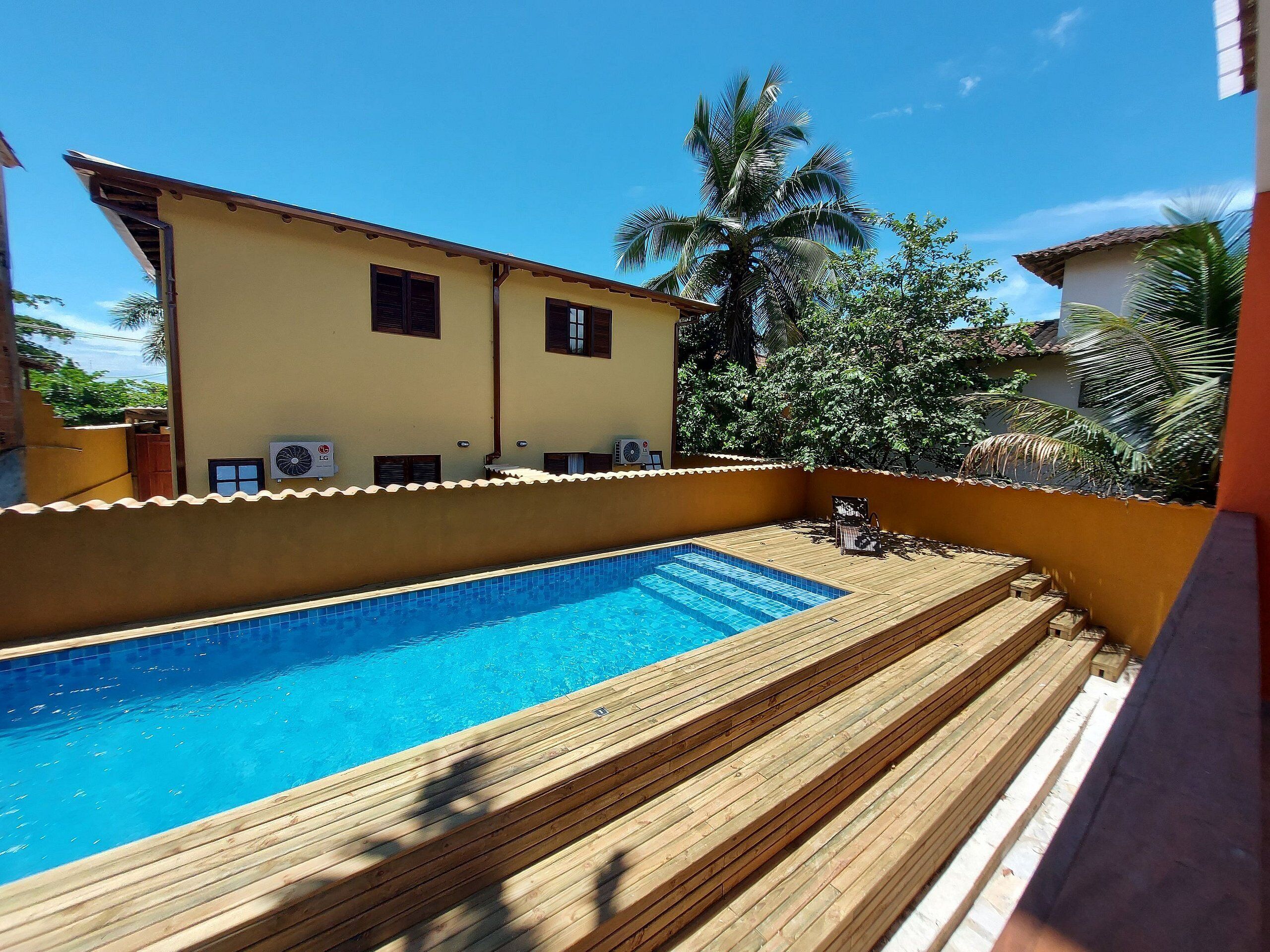 Une piscine avec une terrasse en bois, un bâtiment jaune doté de climatiseurs et, au fond, des palmiers.