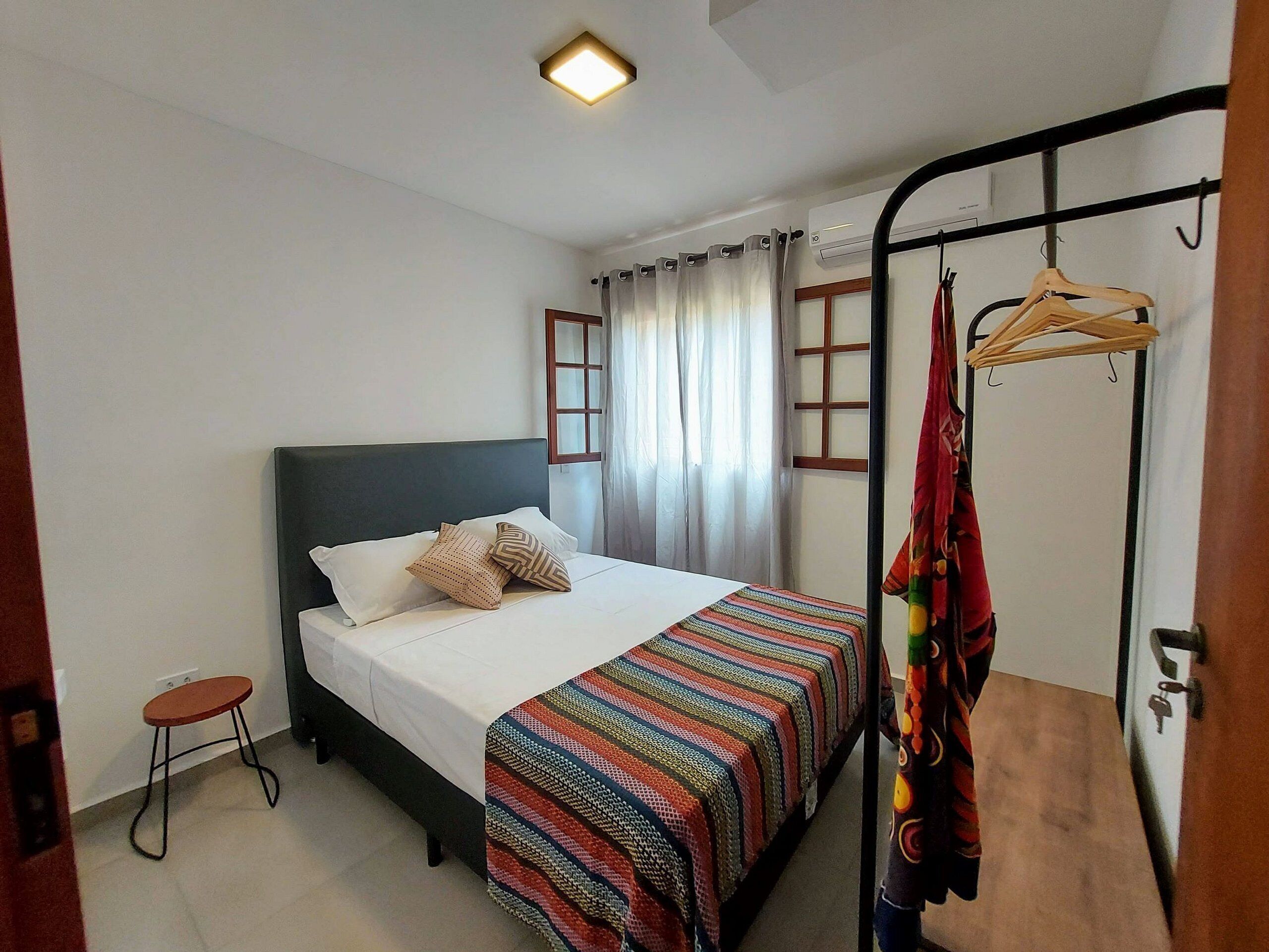 A bedroom with a bed, a small stool, a hanging clothes rack, and a window with curtains.