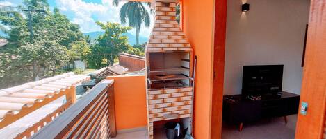 A balcony with a brick barbecue grill, a TV, and a view of greenery and mountains.