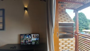 A room with a TV displaying various channels, a brick oven, and a view of the outdoors. - Cepilho - Cosy Flat Near the Beach with kitchen, private BBQ, pool (Paraty)