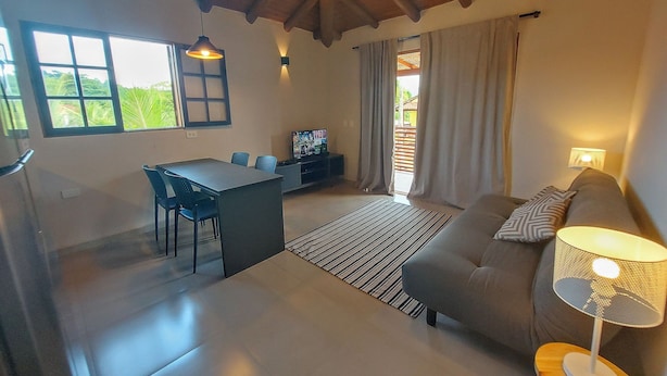 A modern living room with a sofa, a dining table with chairs, a TV, and a lamp. - Cepilho - Cosy Flat Near the Beach with kitchen, private BBQ, pool (Paraty)