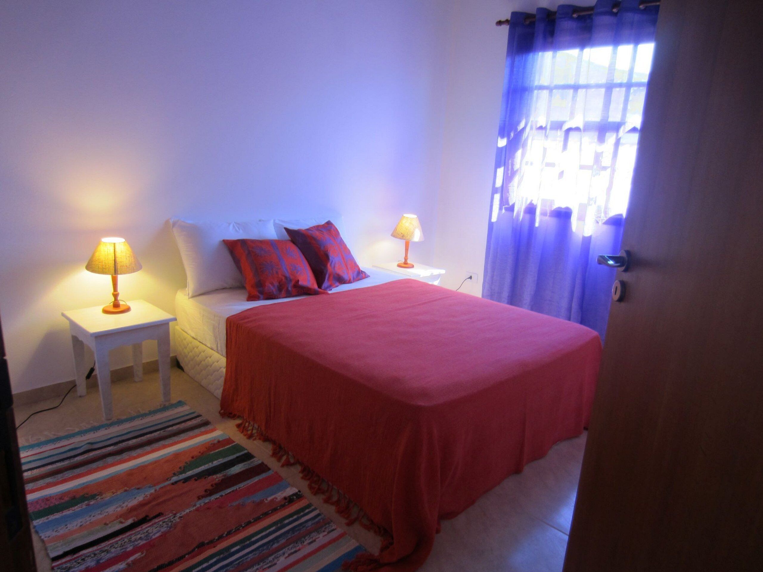 Une chambre à coucher avec un lit, deux tables de chevet avec des lampes, un tapis rayé aux couleurs vives et une fenêtre avec des rideaux.