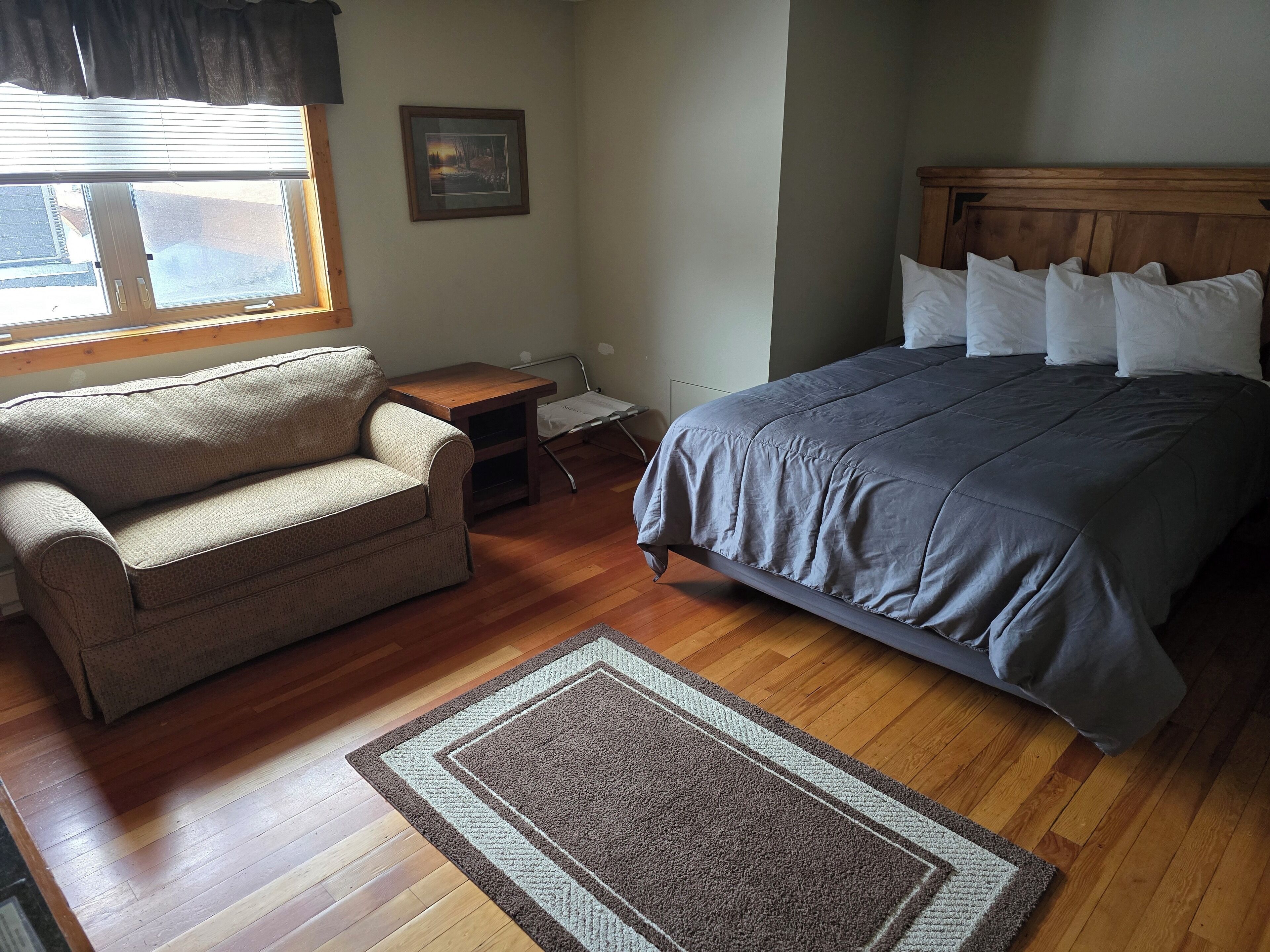 A bedroom with a bed, a sofa, a wooden nightstand, a framed picture on the wall, and a window with blinds.