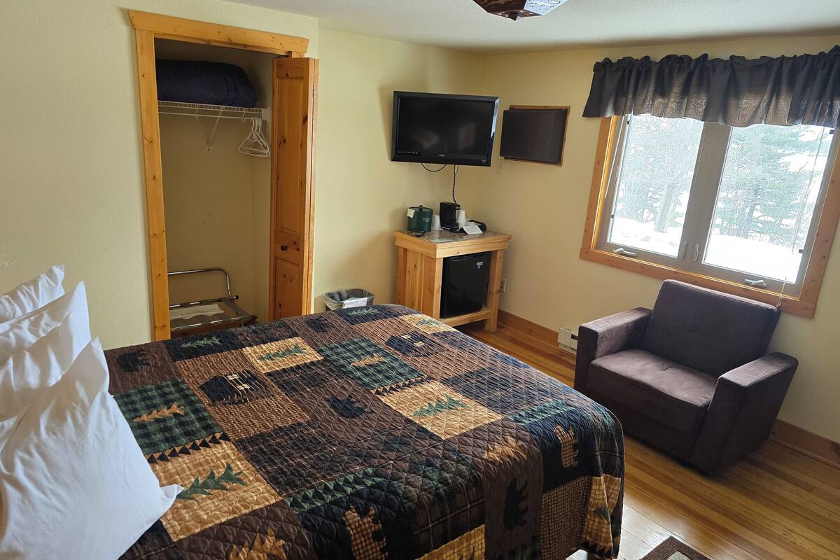 Un dormitorio con cama, una silla, un escritorio con computadora, televisión y ventana con vista a árboles.
