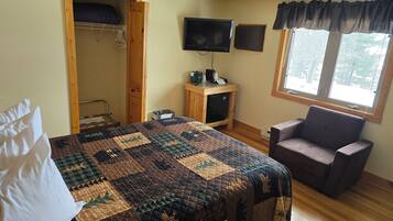 A bedroom with a bed, a chair, a desk with a computer, a TV, and a window with a view of trees.
