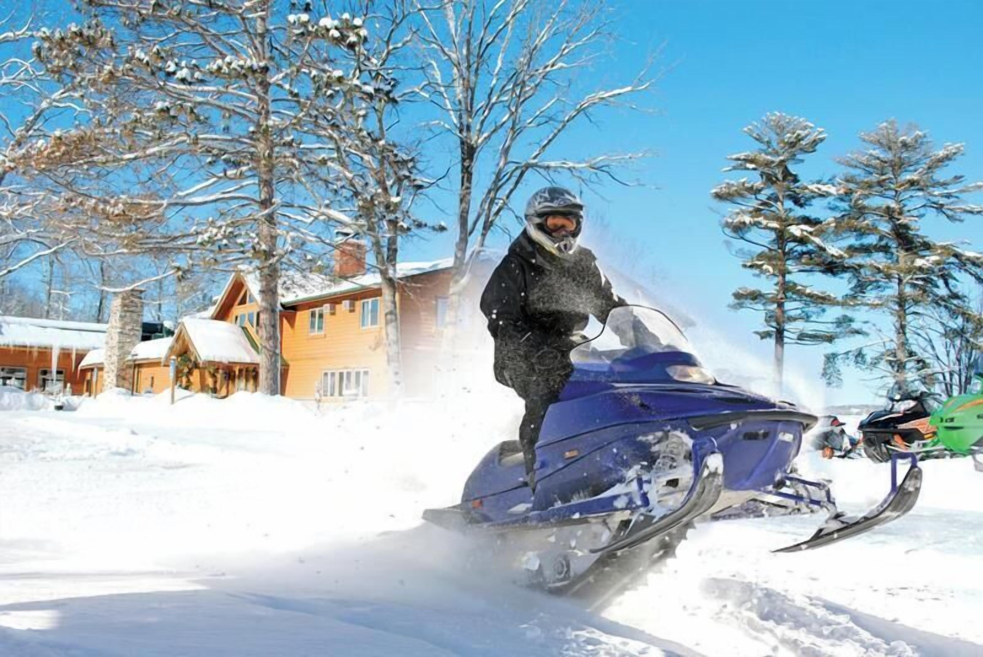 Un vehículo para nieve circulando por un camino nevado con casas al fondo.