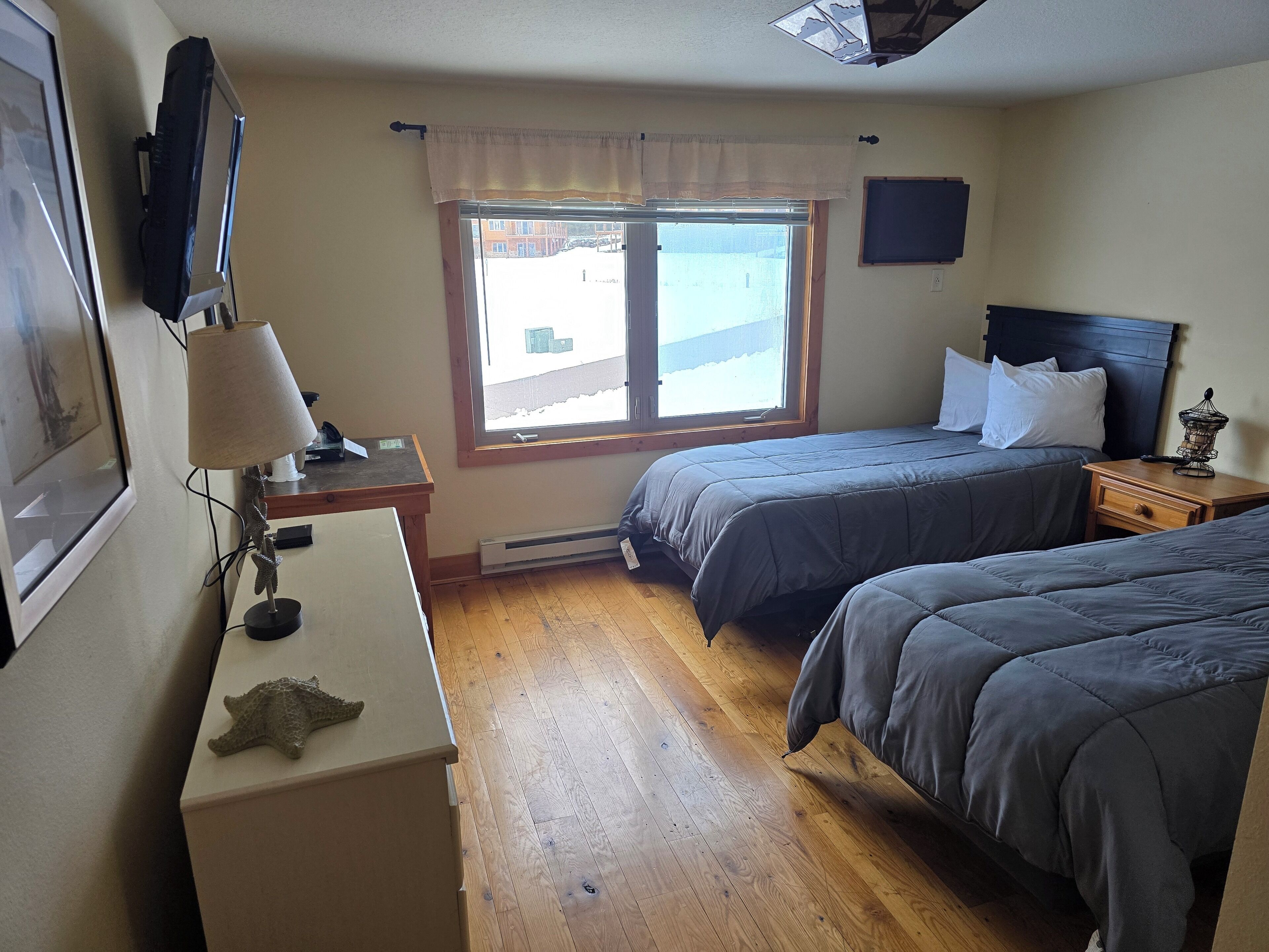 A bedroom with two beds, a desk, a lamp, and a window with a view of buildings.