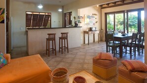 Amplia sala de estar con cocina, mesa y sillas de comedor. Un sofá y un reposapiés se ven en primer plano, y un ventanal ofrece vistas al exterior.