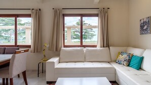 A modern living room with a white sofa, a dining table, and a chair. There are large windows with curtains, a lamp, and a painting on the wall. - Safira House. Beautiful 3 suite house in front of the sea with private swimming pool and BBQ area (Paraty)