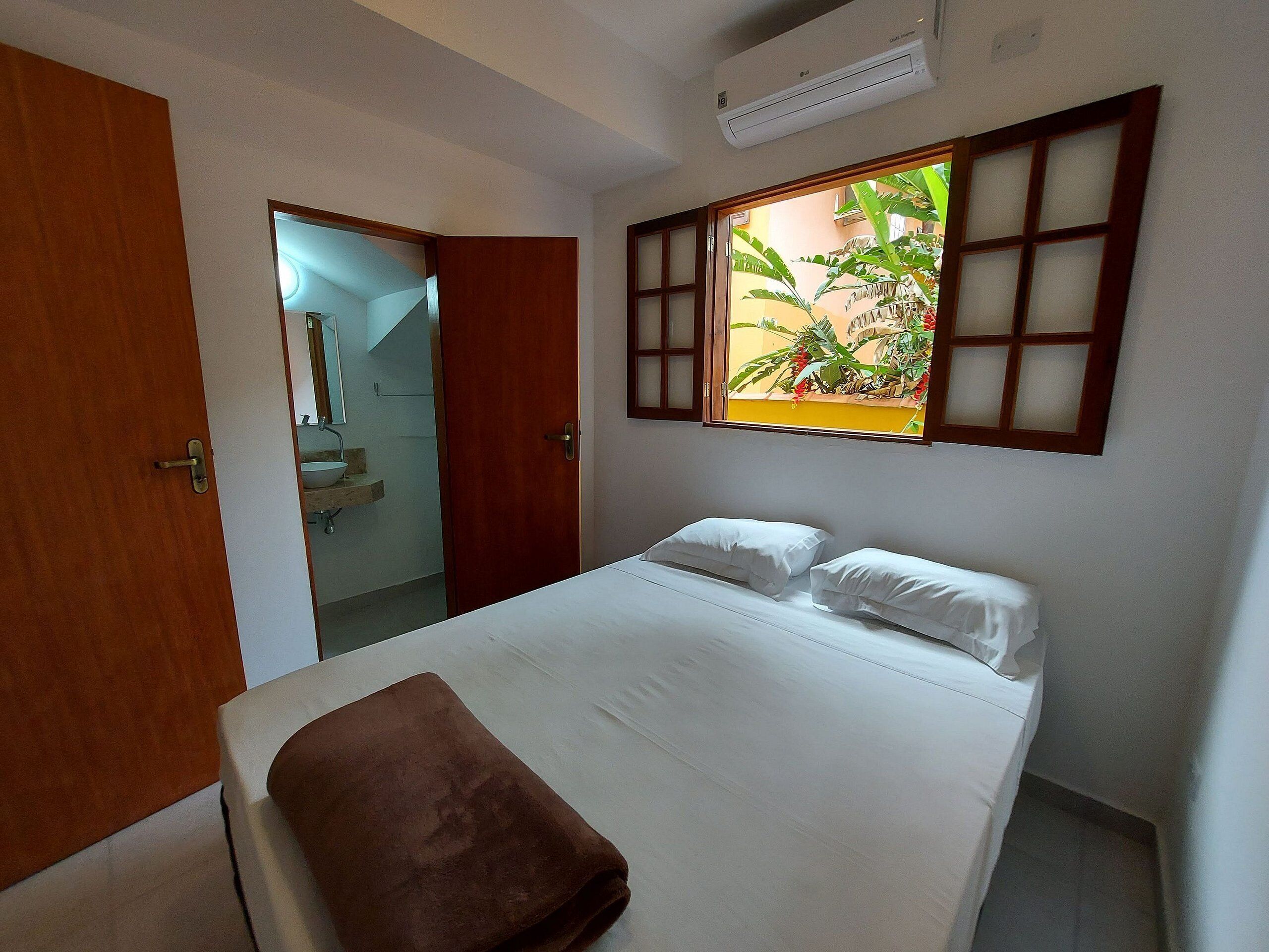 Une chambre à coucher avec un lit, une fenêtre donnant sur des plantes, une salle de bain avec un lavabo et un appareil de climatisation.