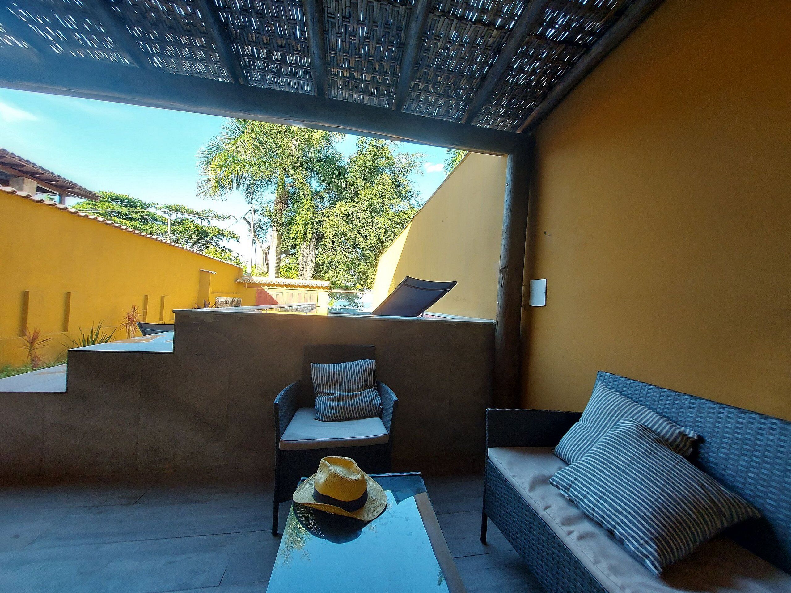 A covered outdoor seating area with a sofa and chairs, a table with a hat, and a view of a yellow building and trees.