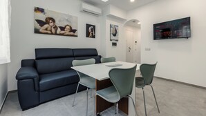 Un salon moderne avec un canapé noir, une table blanche et des chaises vertes. On y trouve des tableaux accrochés au mur, un téléviseur et un appareil de climatisation.