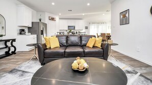 Una sala de estar moderna con un sofá de cuero negro, una mesa de centro con productos de panadería y una cocina al fondo.