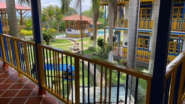 Vista desde un balcón con suelo de baldosas, que da a un área de piscina con palmeras y un edificio con detalles en amarillo y azul.