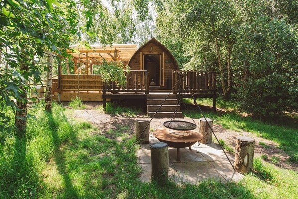 Una cabaña rústica con terraza de madera, un brasero y una zona de estar al aire libre cubierta, rodeada de árboles.