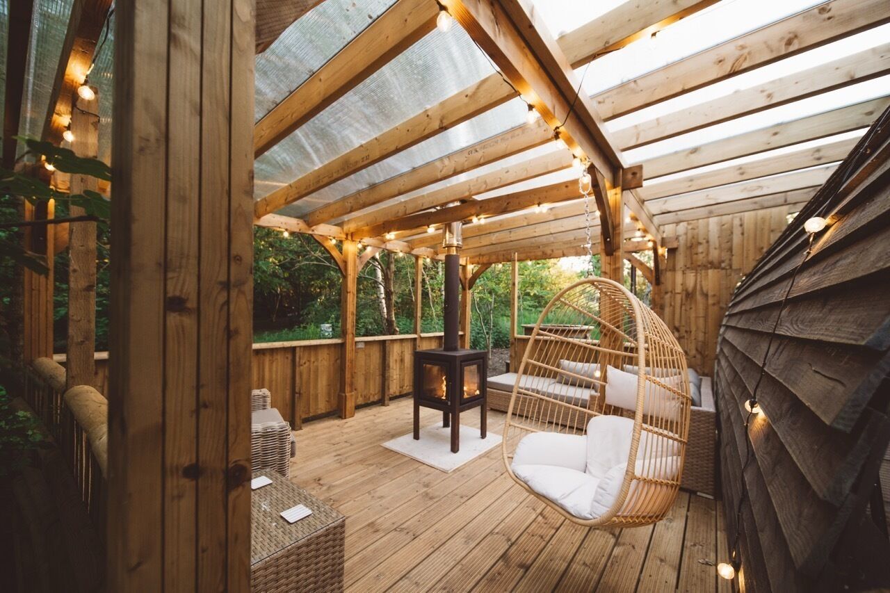 Una terraza de madera con techo de vidrio, una silla colgante y un estufa de leña.