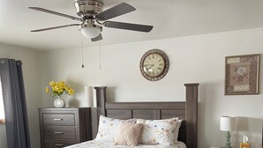 Une chambre à coucher comprenant un lit, une table de chevet, une commode, un ventilateur de plafond, une horloge et un vase à fleurs.