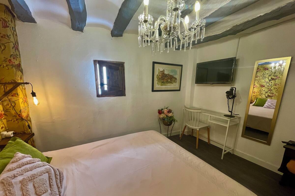 A bedroom with a large bed, a desk, a chair, a mirror, and a chandelier.