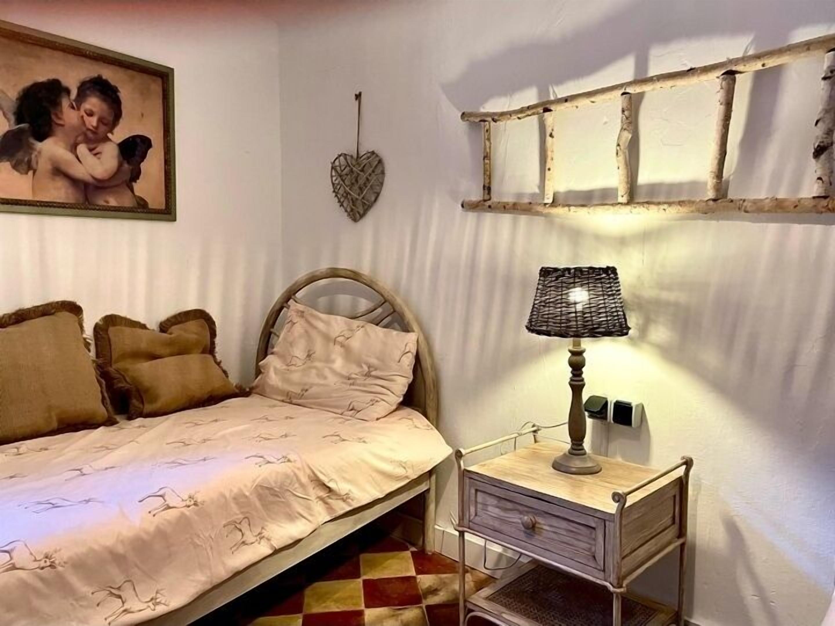 A bedroom with a bed, a bedside table with a lamp, a wall-mounted shelf, and a painting.
