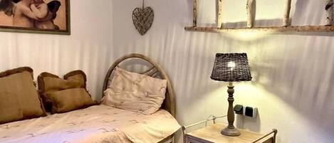 A bedroom with a bed, a bedside table with a lamp, a wall-mounted shelf, and a painting.